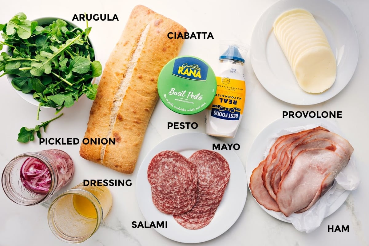 Ingredients In This Ciabatta Sandwich All the ingredients in this recipe prepped out for easy assembly including the arugula, pickled onions, dressing, ciabatta, pesto, mayo, salami, ham, and provolone.