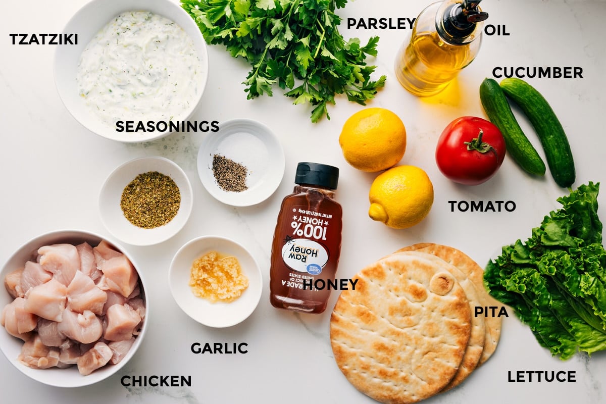 Ingredients In Chicken Souvlaki All the ingredients in this recipe prepped out for easy assembly including the tzatziki, seasonings, chicken, garlic, honey, pita, lettuce, tomato, cucumber, lemon, oil, parsley.