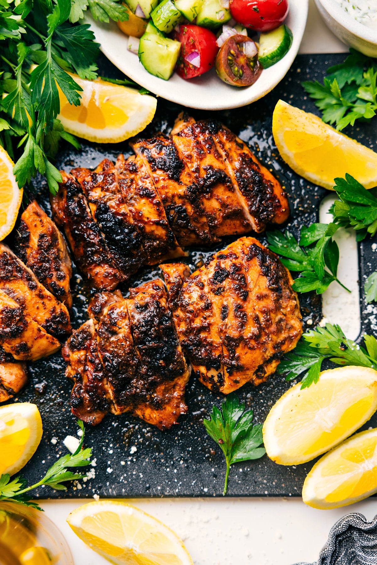 Greek Chicken Thighs Fresh Out Of The Oven Greek Chicken Thighs on a cutting board with fresh lemons and parsley on the side.