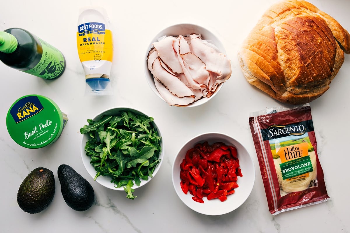 All the ingredients in this recipe prepped out for easy assembly including the oil, pesto, avocado, arugula, turkey, roasted red peppers, provolone, and sourdough.