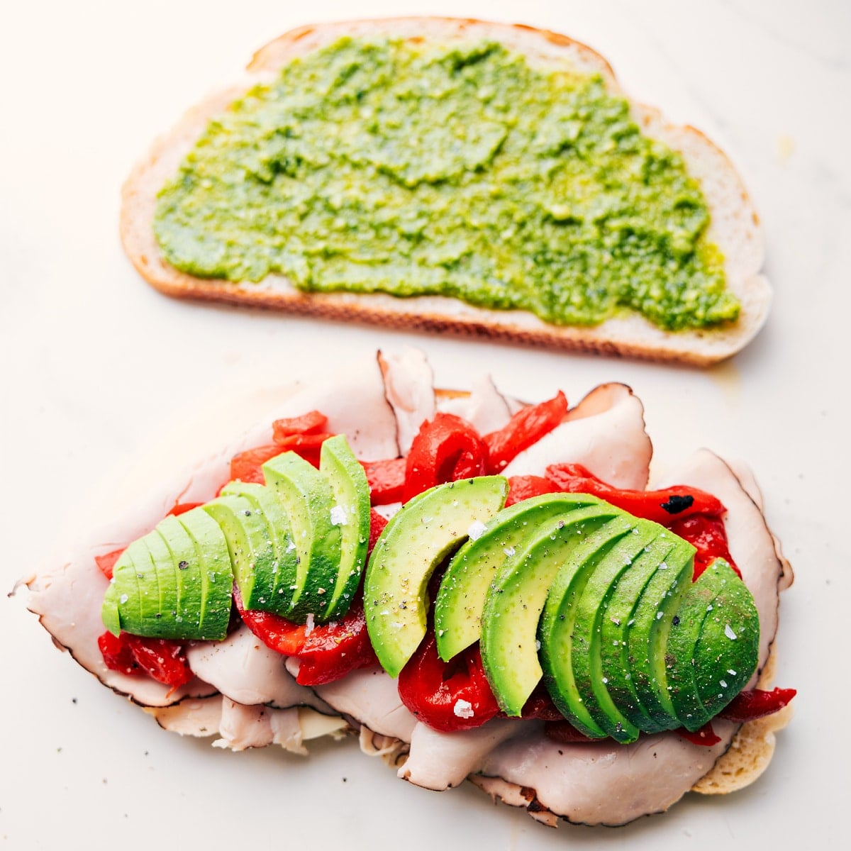 Preparing Panini Sandwich Recipes by layering turkey, cheese, avocado, roasted peppers, arugula, and pesto onto sliced bread.