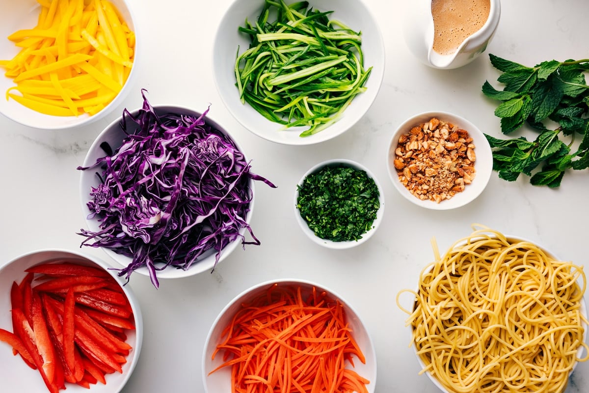All the ingredients in this recipe prepped out for easy assembly including the peppers, cabbage, carrots, pasta, peanuts, dressing, herbs, and cucumbers.