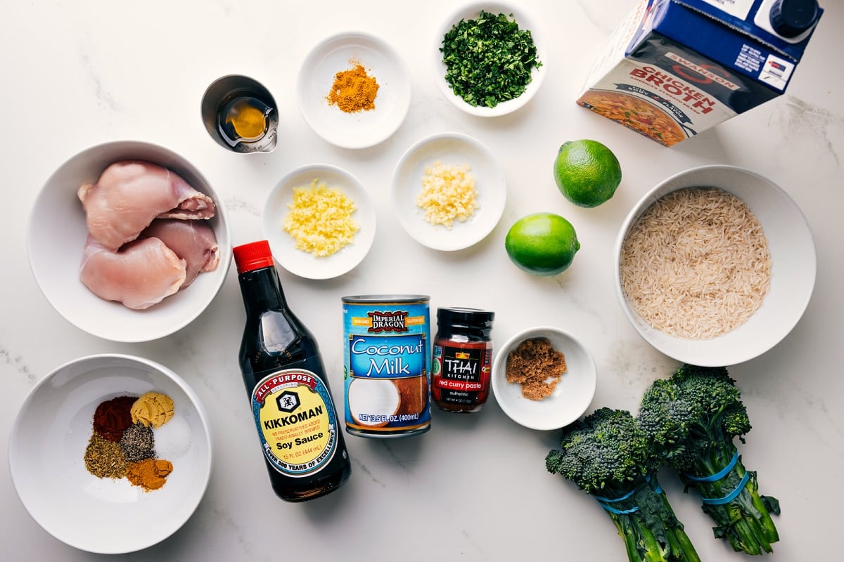 All the ingredients in this recipe prepped out for easy assembly including the chicken thighs, seasonings, garlic, ginger, soy sauce, coconut milk, curry paste, broccoli, rice, limes, herbs, and broth.