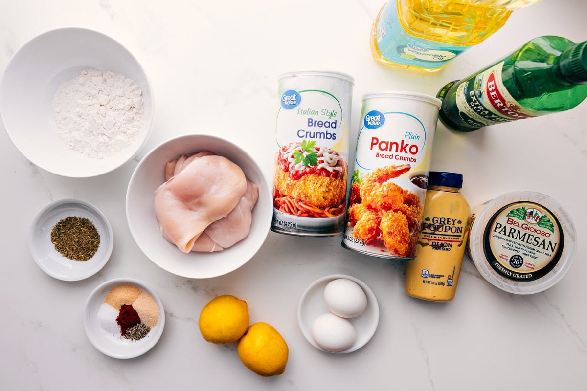 All the ingredients in this recipe prepped out for easy assembly including the flour, seasonings, chicken, lemon, egg, bread crumbs, dijon, parmesan, and oil.