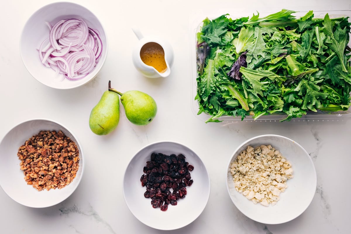 Pear Salad starts with mixed greens lettuce Ingredients in this recipe prepped out for easy assembly including the onions, candied walnuts, cranberries, gorgonzola, mixed greens, dressing, and pears.