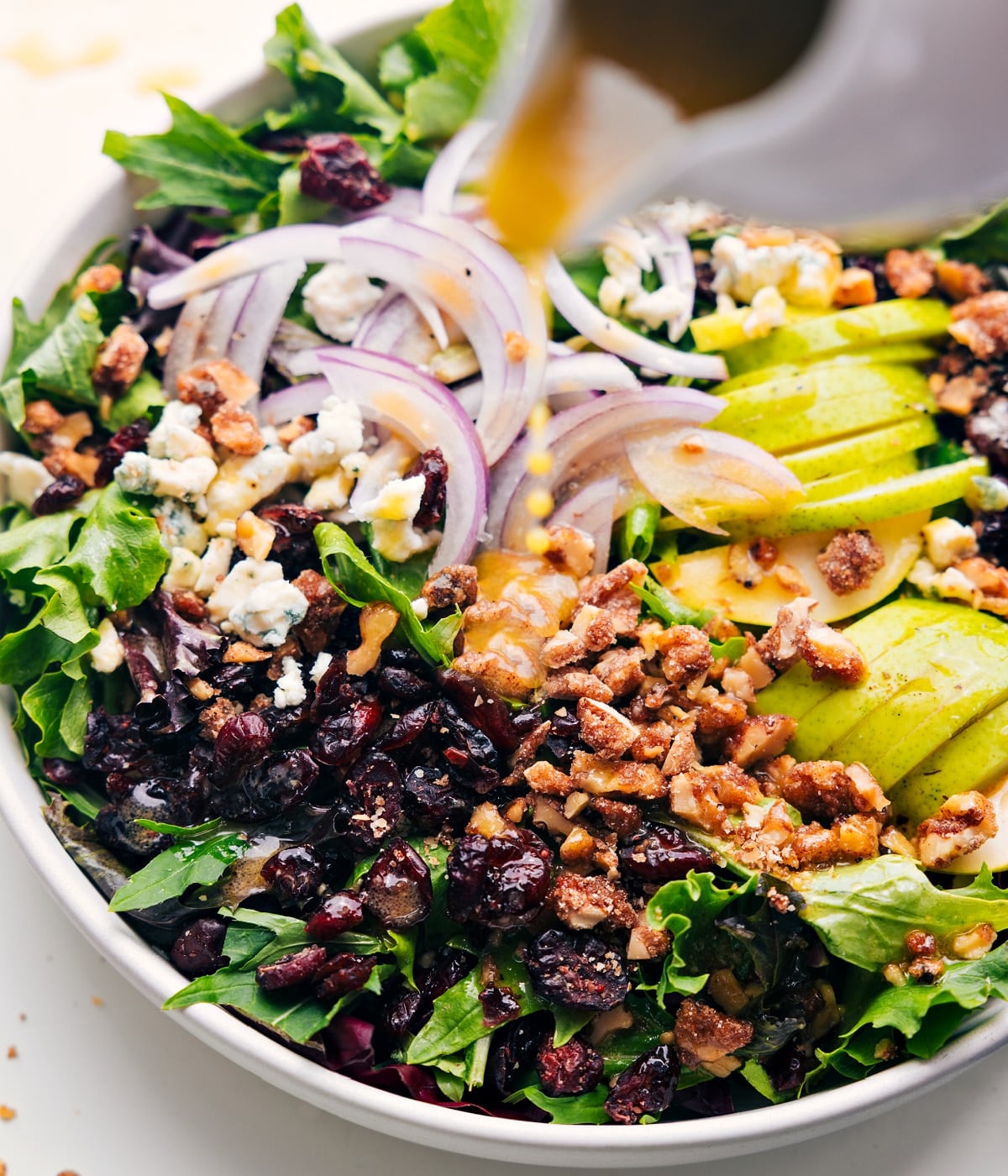 Pear Salad starts with mixed greens lettuce and is loaded with juicy, ripe pears, sweet dried cranberries, candied walnuts, tart red onion, and gorgonzola cheese. The dressing being poured over the salad.