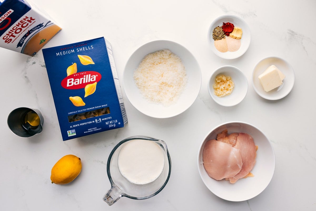 All the ingredients in this recipe prepped out for easy assembly including the stock, shells, oil, lemon, heavy cream, parmesan, chicken, seasonings, garlic, and butter.