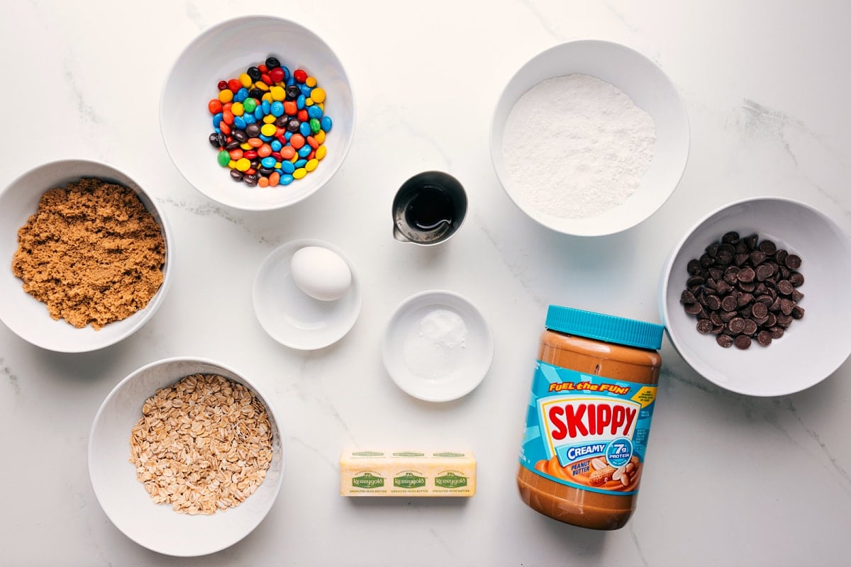 All the ingredients in this recipe prepped out for easy assembly including the sugar, oats, butter, egg, baking soda, salt, peanut butter, chocolate chips, flour, vanilla, and M&Ms.