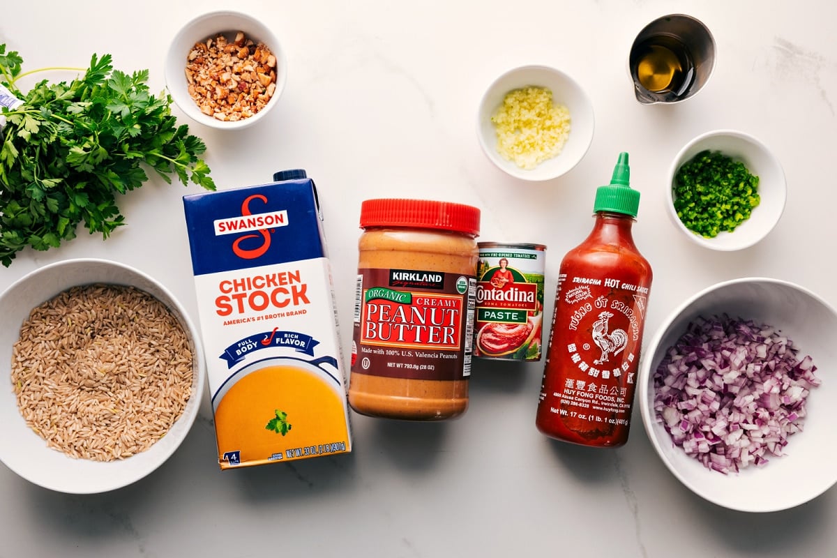 Prepped ingredients for peanut butter soup, including rice, cilantro, stock, peanut butter, paste, sriracha, onion, jalapeño, garlic, peanuts, and oil.
